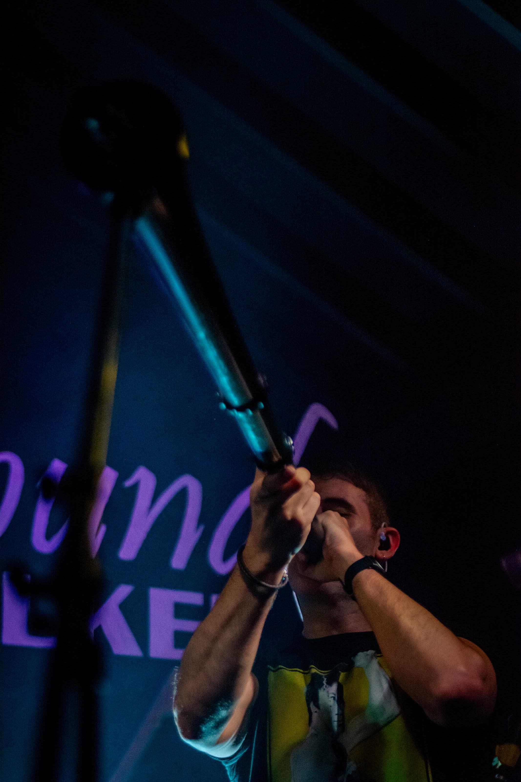 A young man sings into a microphone on a dark stage, a mic stand in the foreground and purple lettering behind him.