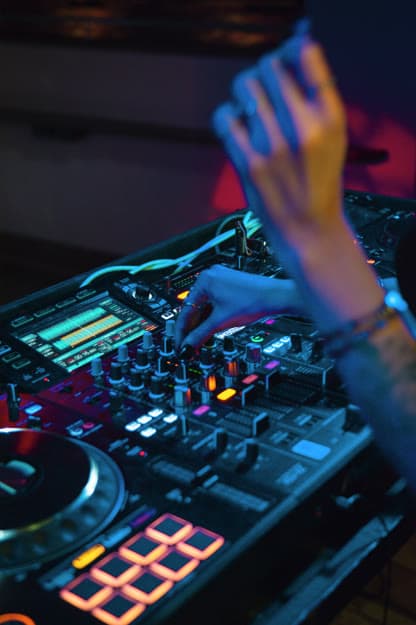 Close-up of hands on a DJ controller, orange performance pads lit, blue and red club light, one hand lifted above the mixer.