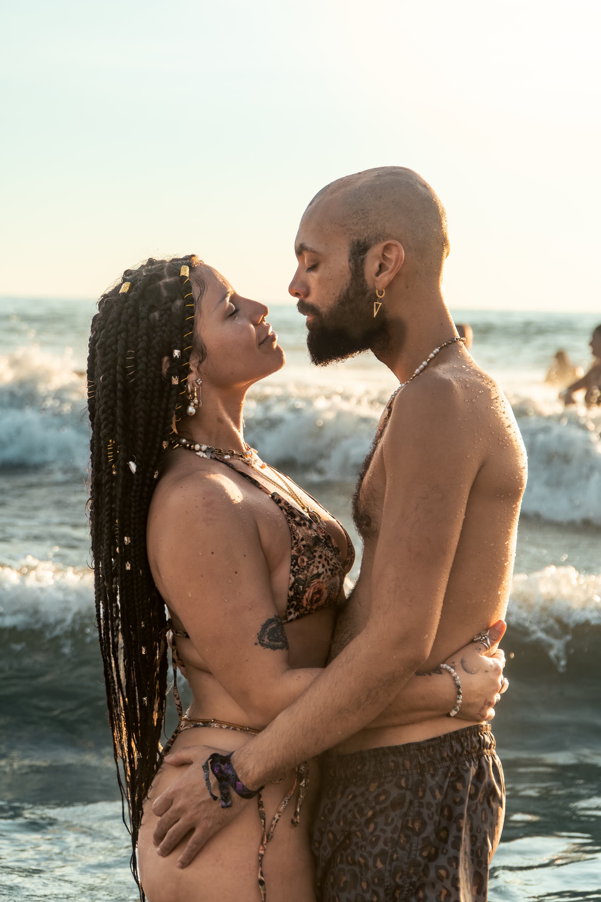 Una pareja metida hasta la cintura en las olas a la hora dorada, ojos cerrados y frentes a punto de tocarse: ella de frente, con trenzas oscuras largas, puños de oro y bikini de leopardo; él calvo y con barba, en bañador de leopardo a juego.