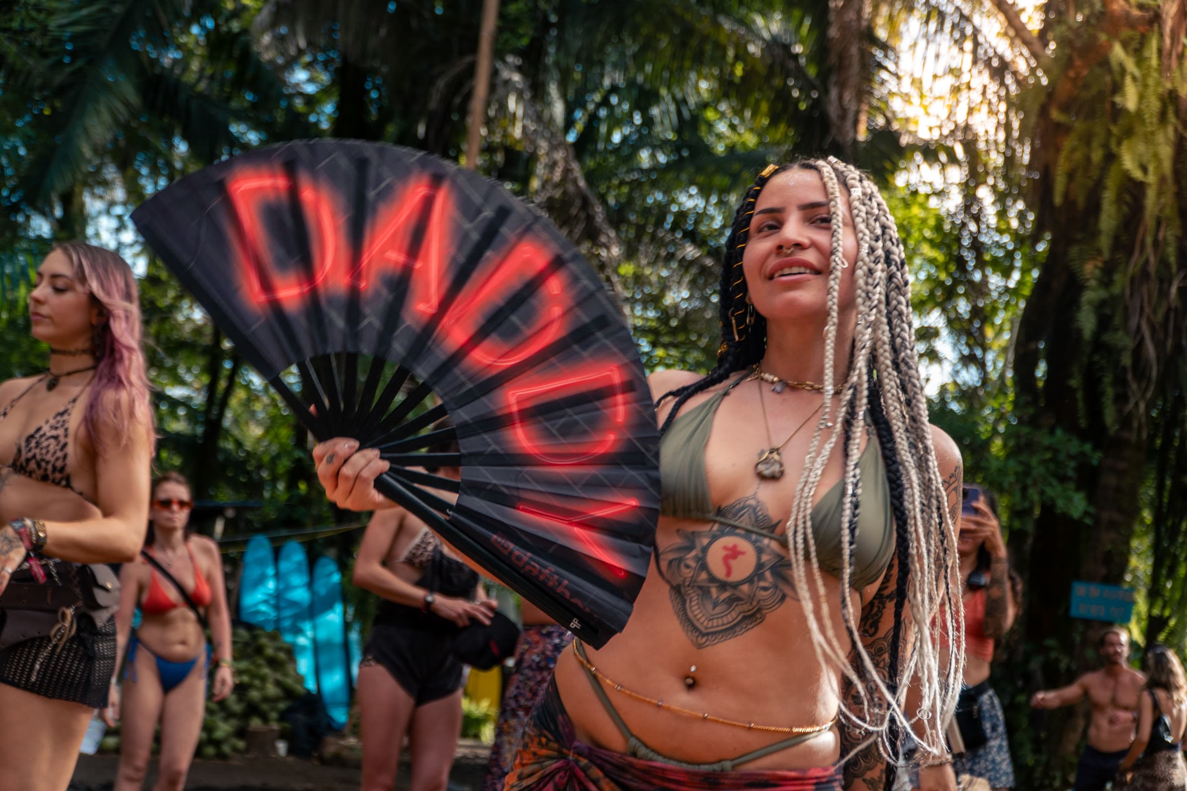 Uma mulher com microtranças loiras e pretas segura um leque vermelho néon onde se lê “DADDY”, com biquíni verde-azeitona e um pareo estampado entre folhagem tropical de festival.