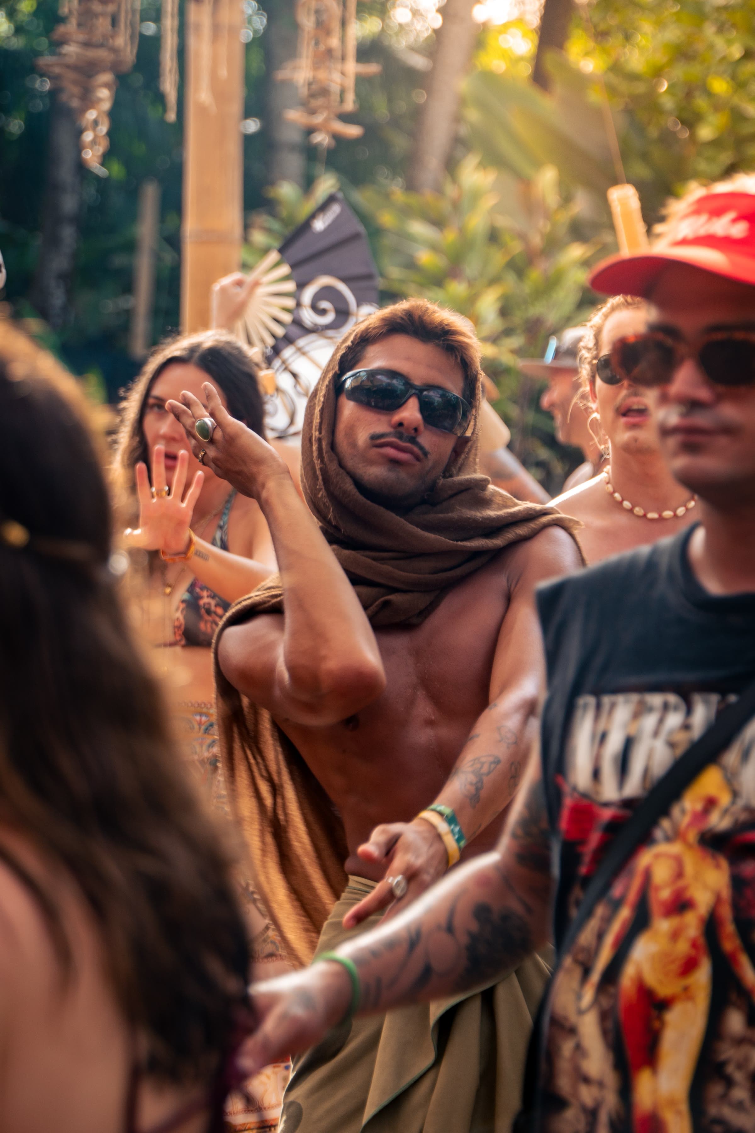 Um homem sem camisa e de óculos de sol dança com um pano castanho sobre a cabeça, com luz dourada e multidão de festival atrás.