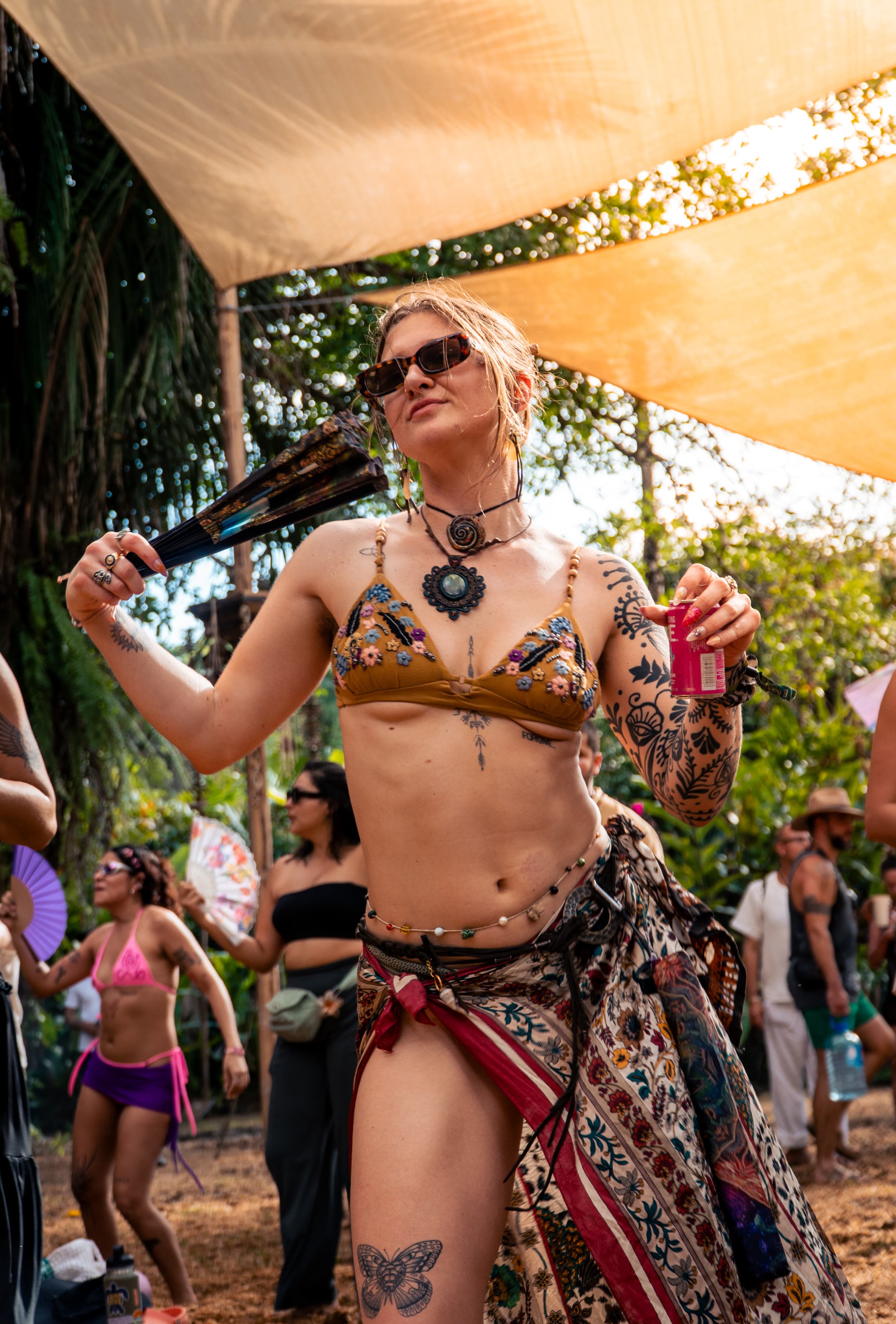 A blonde woman dances under fabric canopies with a patterned fan and a pink drink can, in a gold-embroidered bikini and wrap skirt.