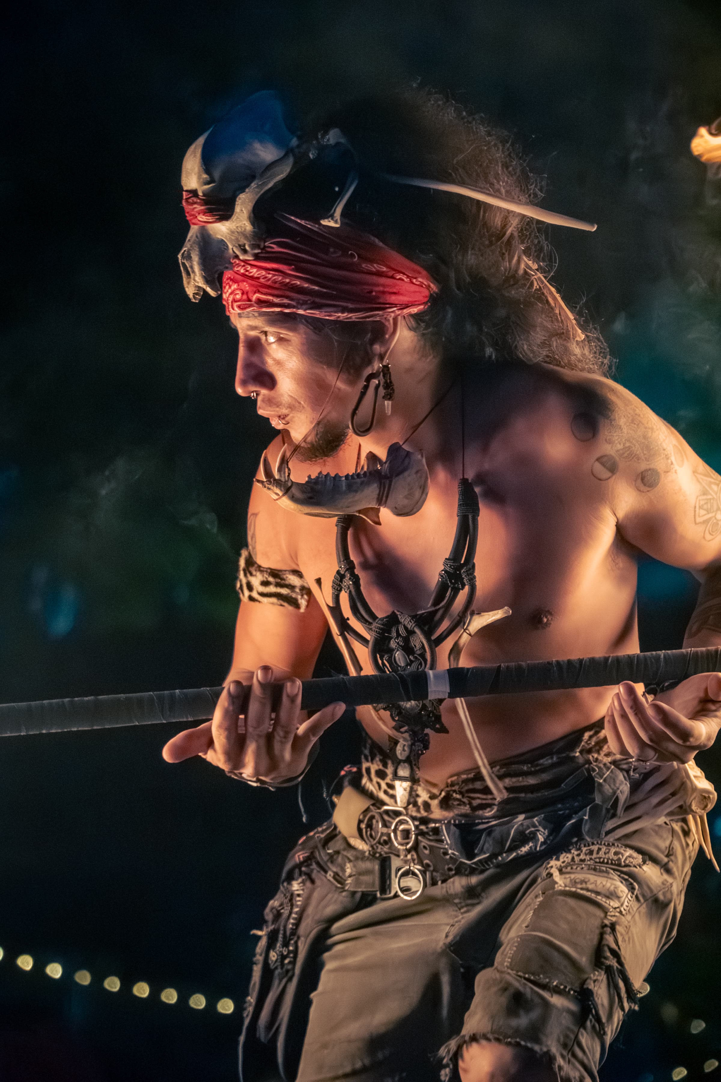 Um homem com um adereço de caveira e lenço vermelho segura um cajado numa performance noturna à luz do fogo.