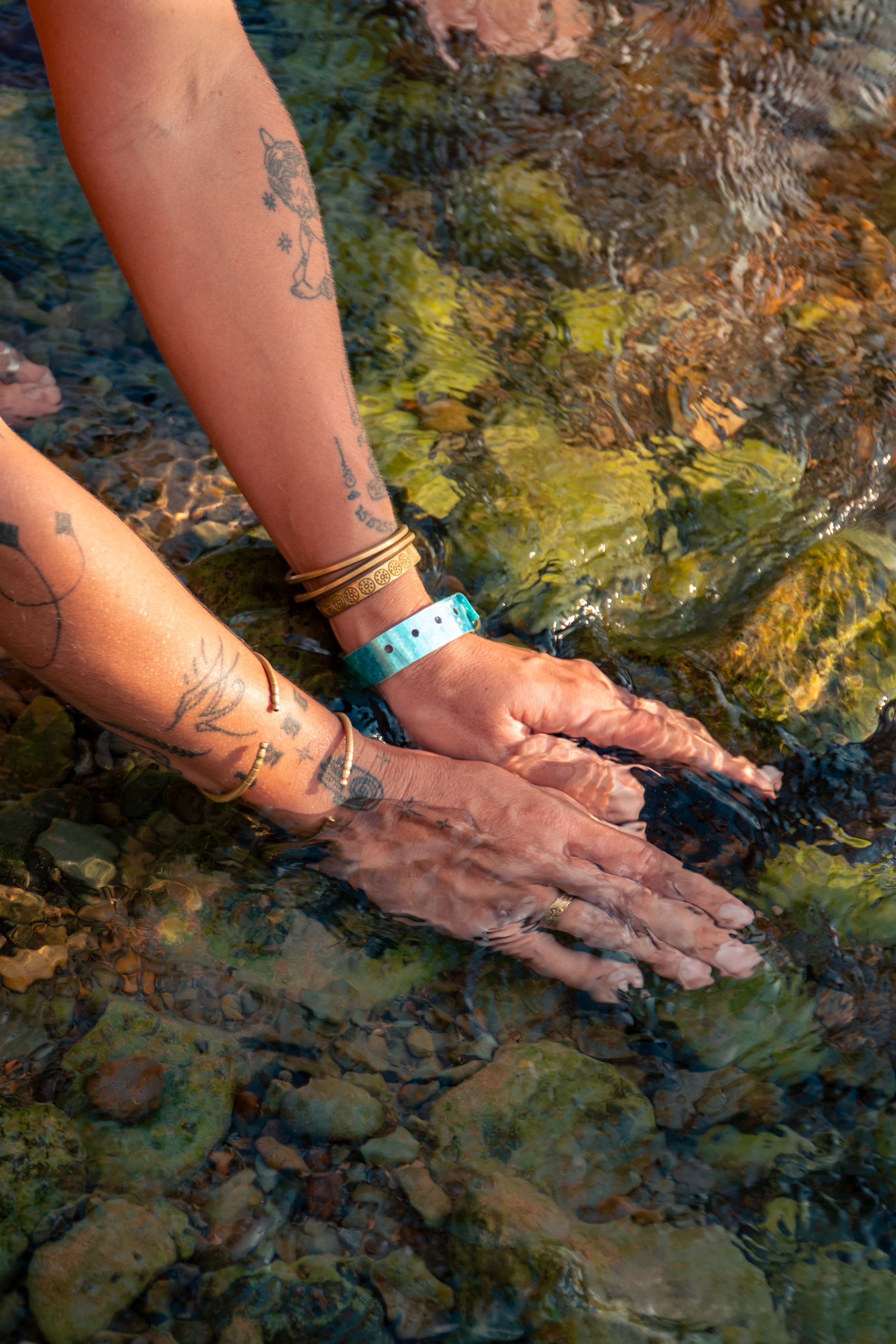 Dois braços tatuados entram em água clara e pouco profunda sobre pedras de rio, com pulseiras de ouro.