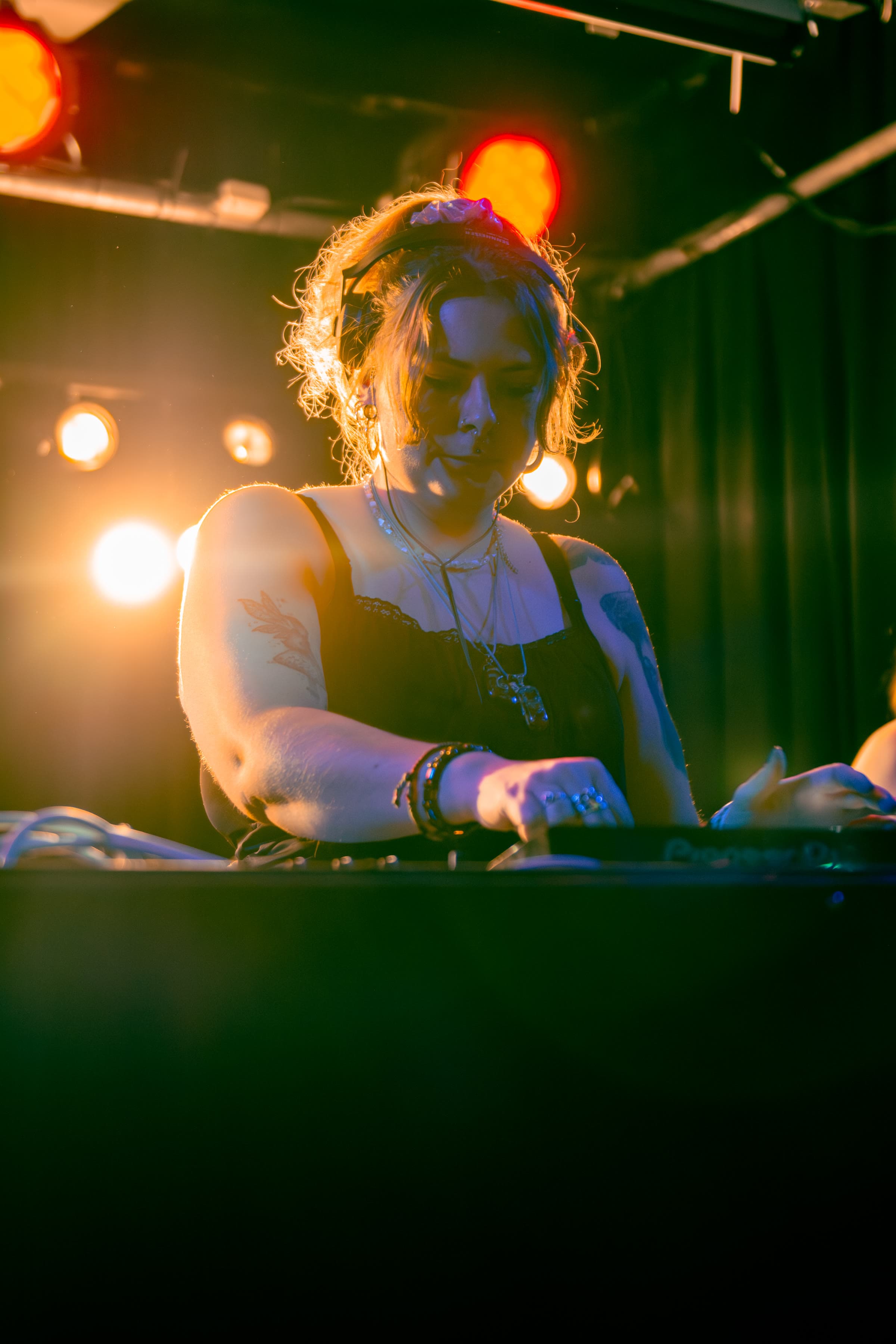 A woman DJ with wavy hair and headphones stands behind a Pioneer deck, warm orange and red stage lights, a lace tank, tattoos along her arm.