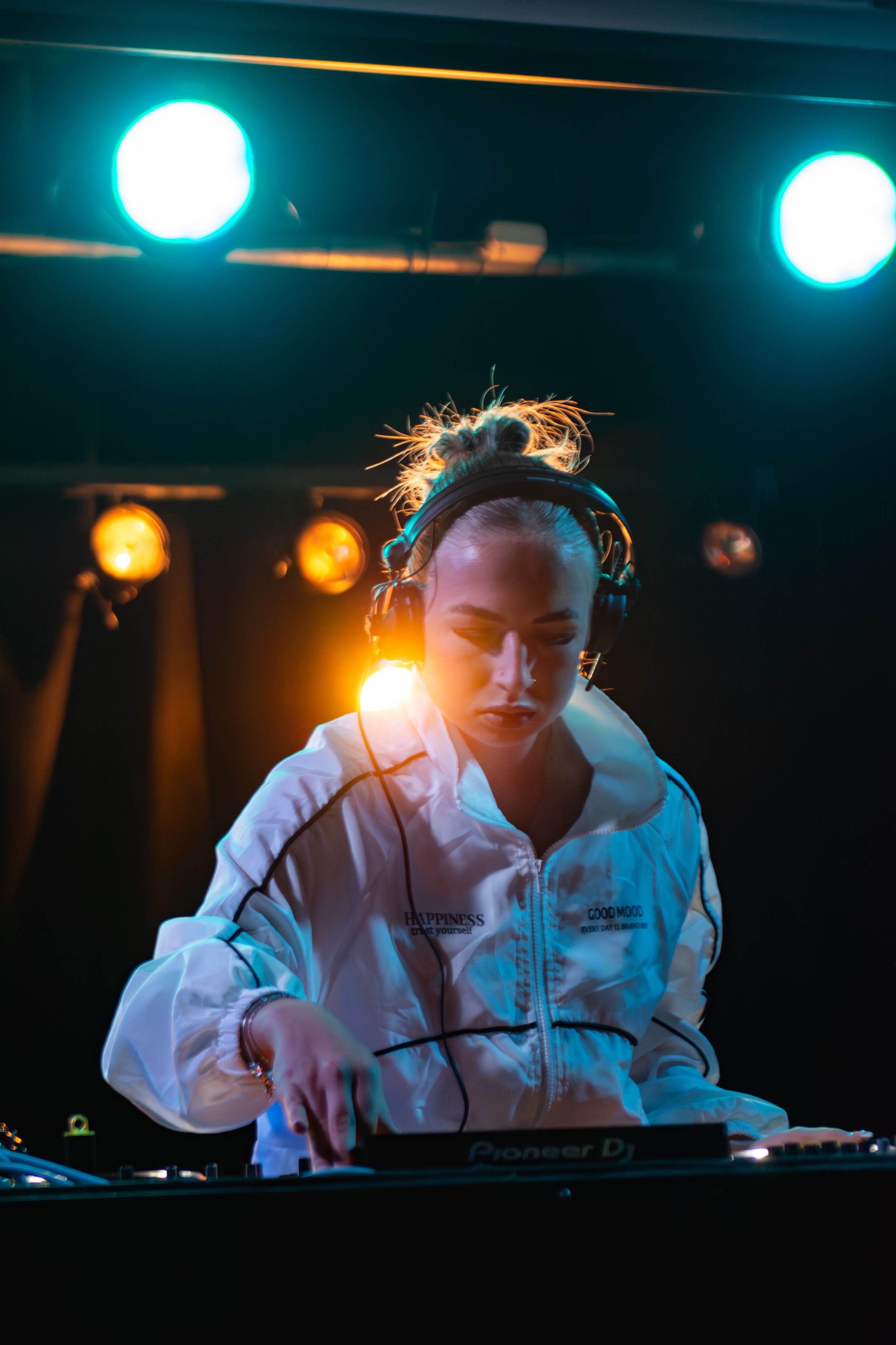 Uma DJ loura com um casaco branco onde se lê “HAPPINESS / GOOD MOOD” e auscultadores, num controlador Pioneer sob luzes ciano e âmbar.