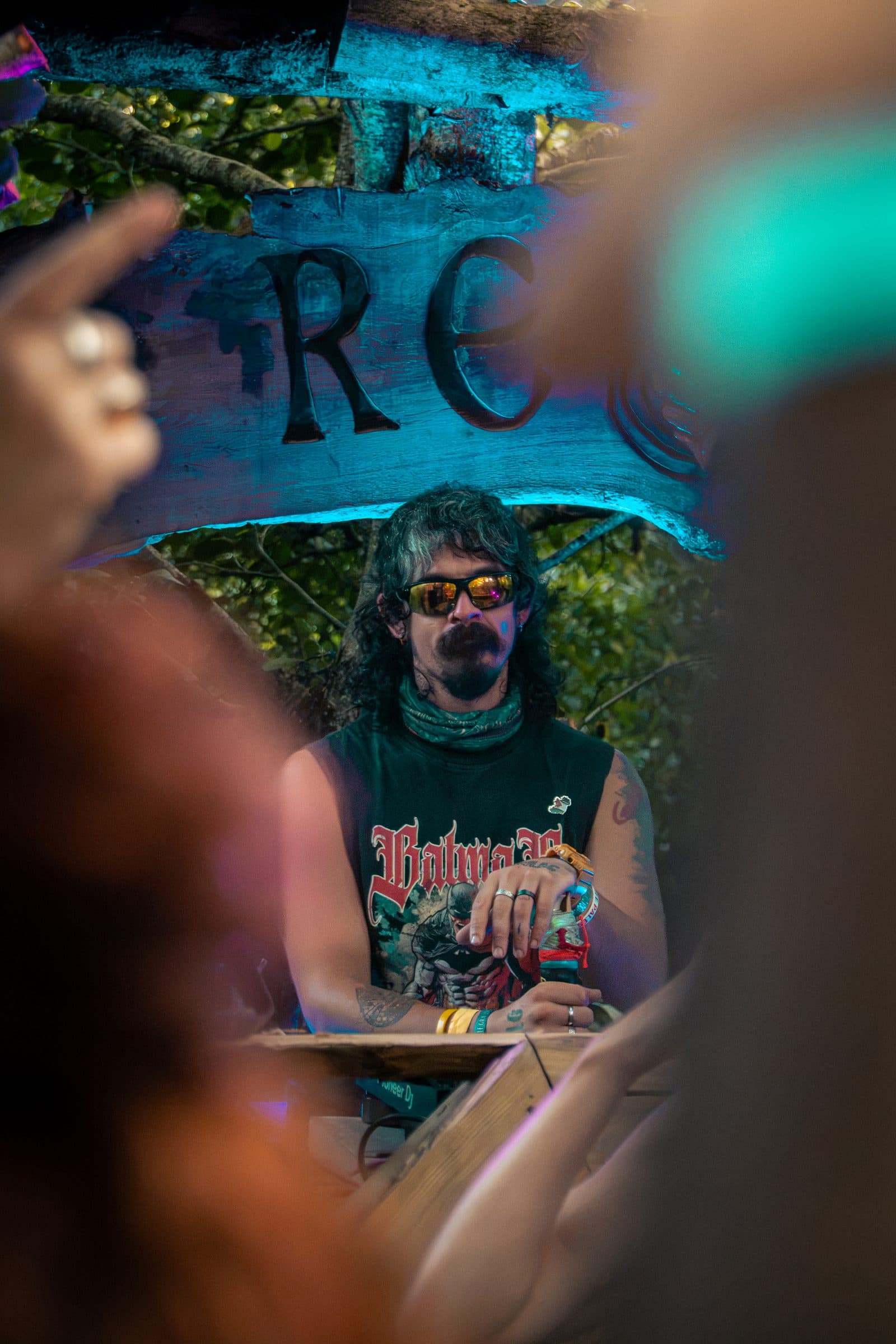 Um homem com cabelo escuro ondulado, bigode e pera, e óculos de lentes amarelas, numa cabine de DJ ao ar livre com uma t-shirt sem mangas verde do Batman, visto através da multidão, um letreiro de madeira “REC” com luz azul.