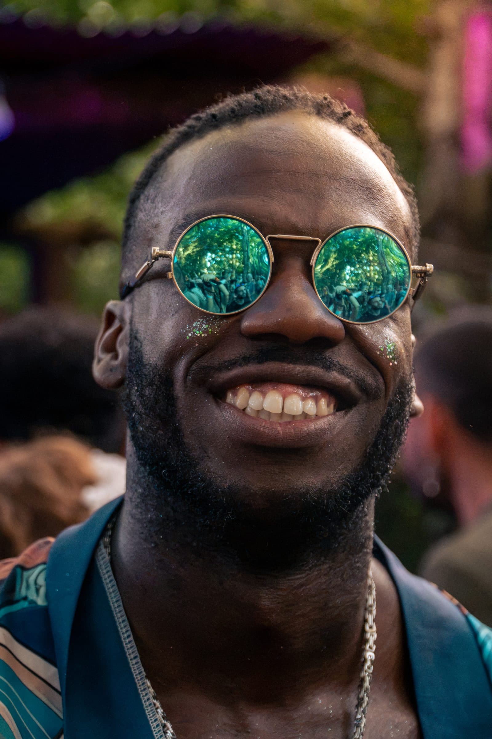 Close-up of a smiling man in round gold-rim mirrored teal sunglasses, cheek glitter, a silver chain and a busy patterned shirt, festival lights soft behind him.