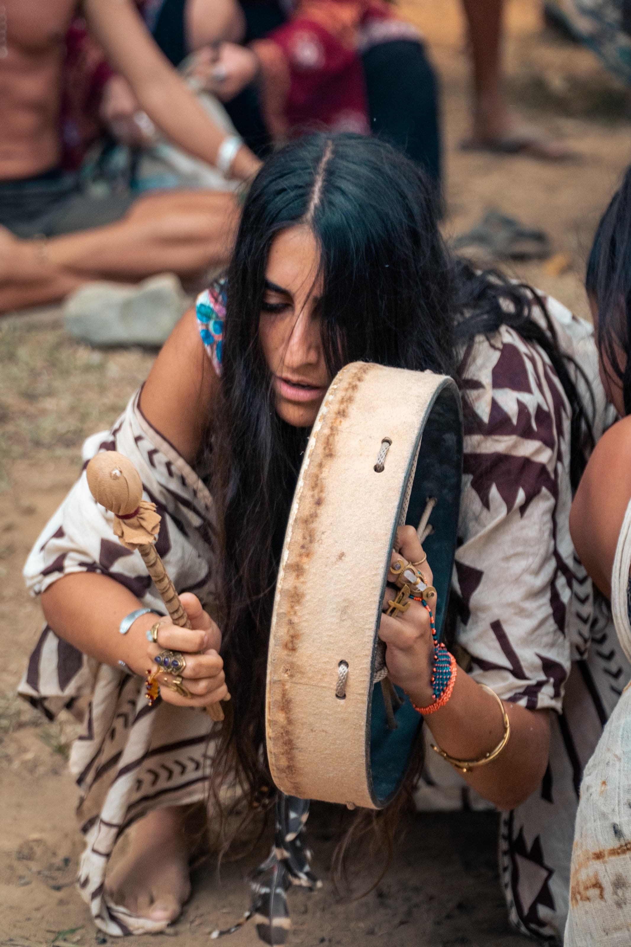 Close-up of a woman at an outdoor gathering holding a large frame drum and a beater, eyes closed, in a blue-green patterned top.