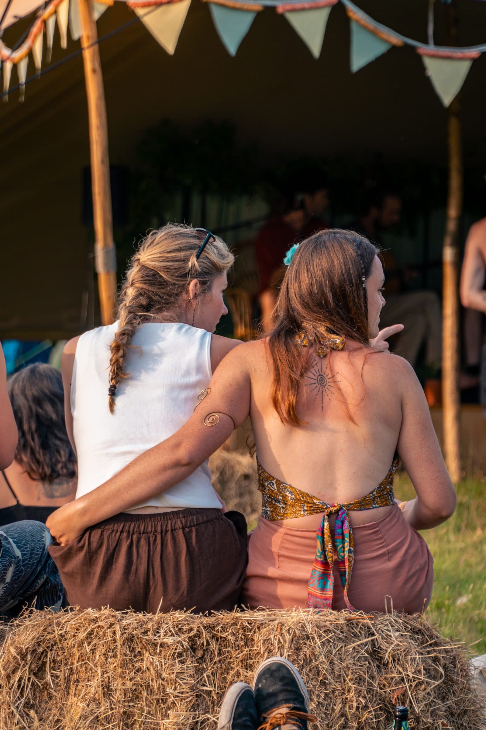 Two women seen from behind sit on a hay bale under a tent strung with colorful bunting: one with a blonde braid and a spiral armlet, the other with dark reddish hair, a backless patterned top, and a sun tattoo on her upper back.