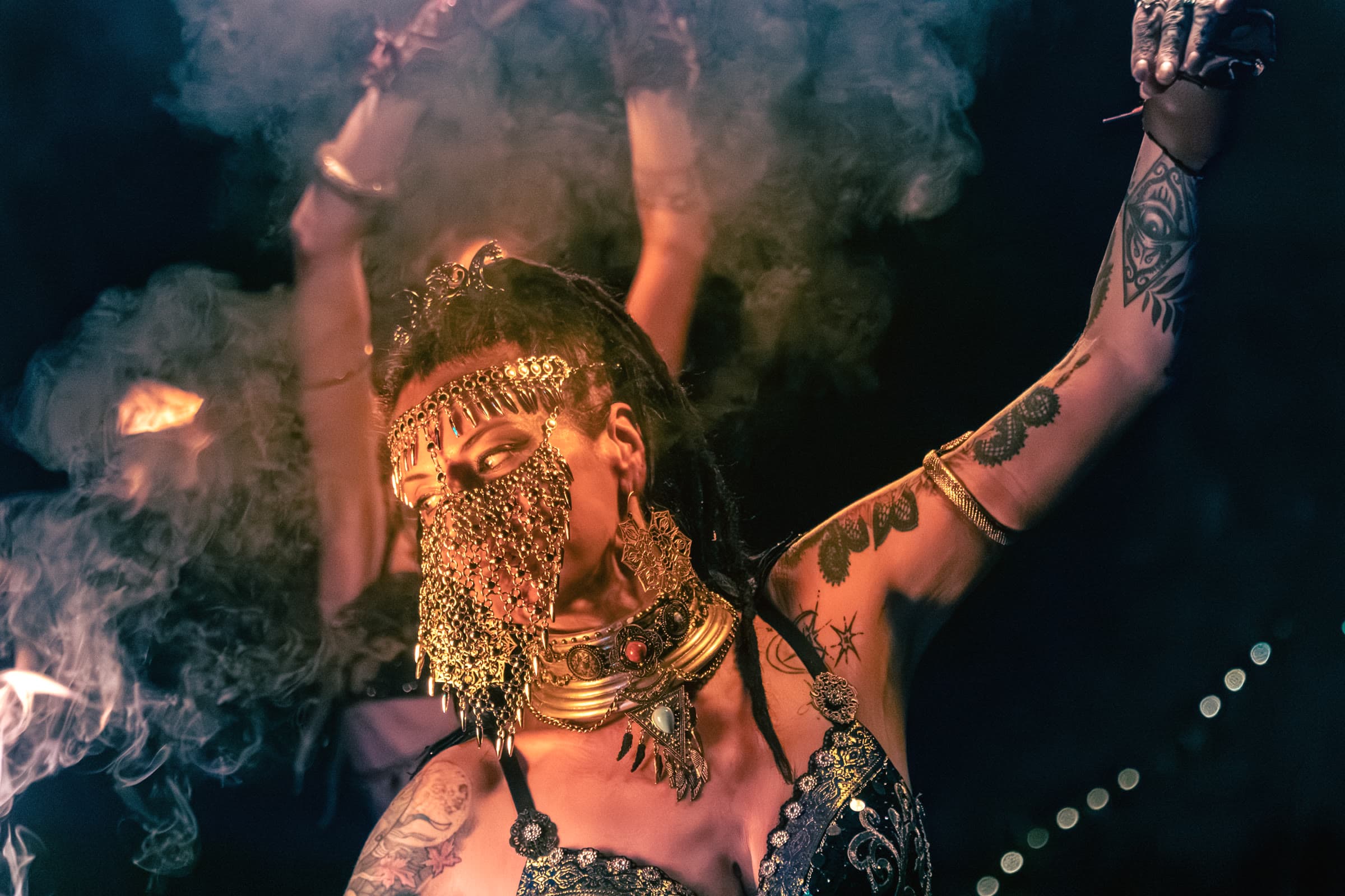 Uma mulher com véu de malha dourada e adereço de lua crescente dança em fumo iluminado pelo fogo, com joalharia tribal-fusion e tatuagens no braço.