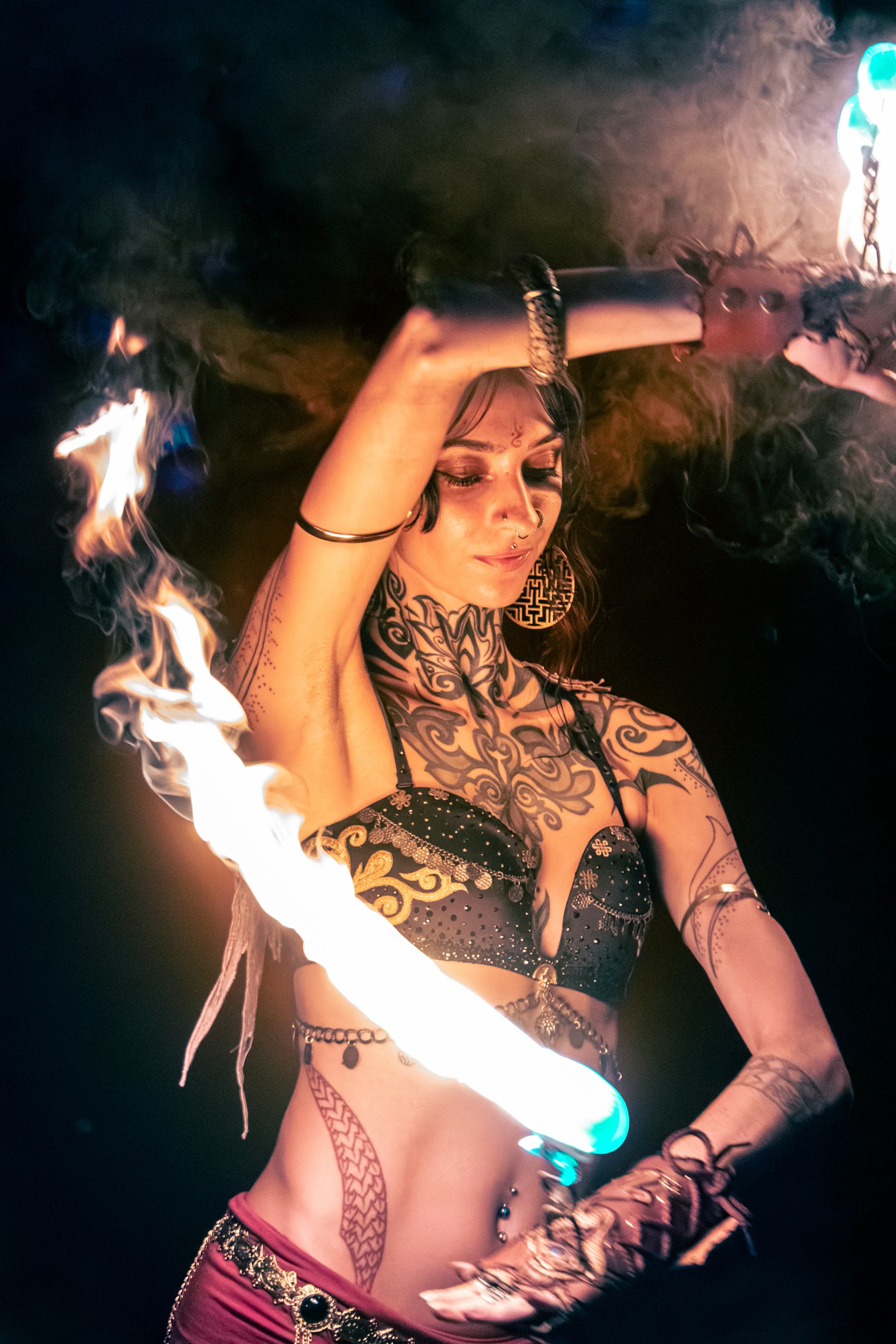 A tattooed fire dancer at night, one arm raised, a long-exposure streak of white-orange flame crossing her torso, smoke behind her.