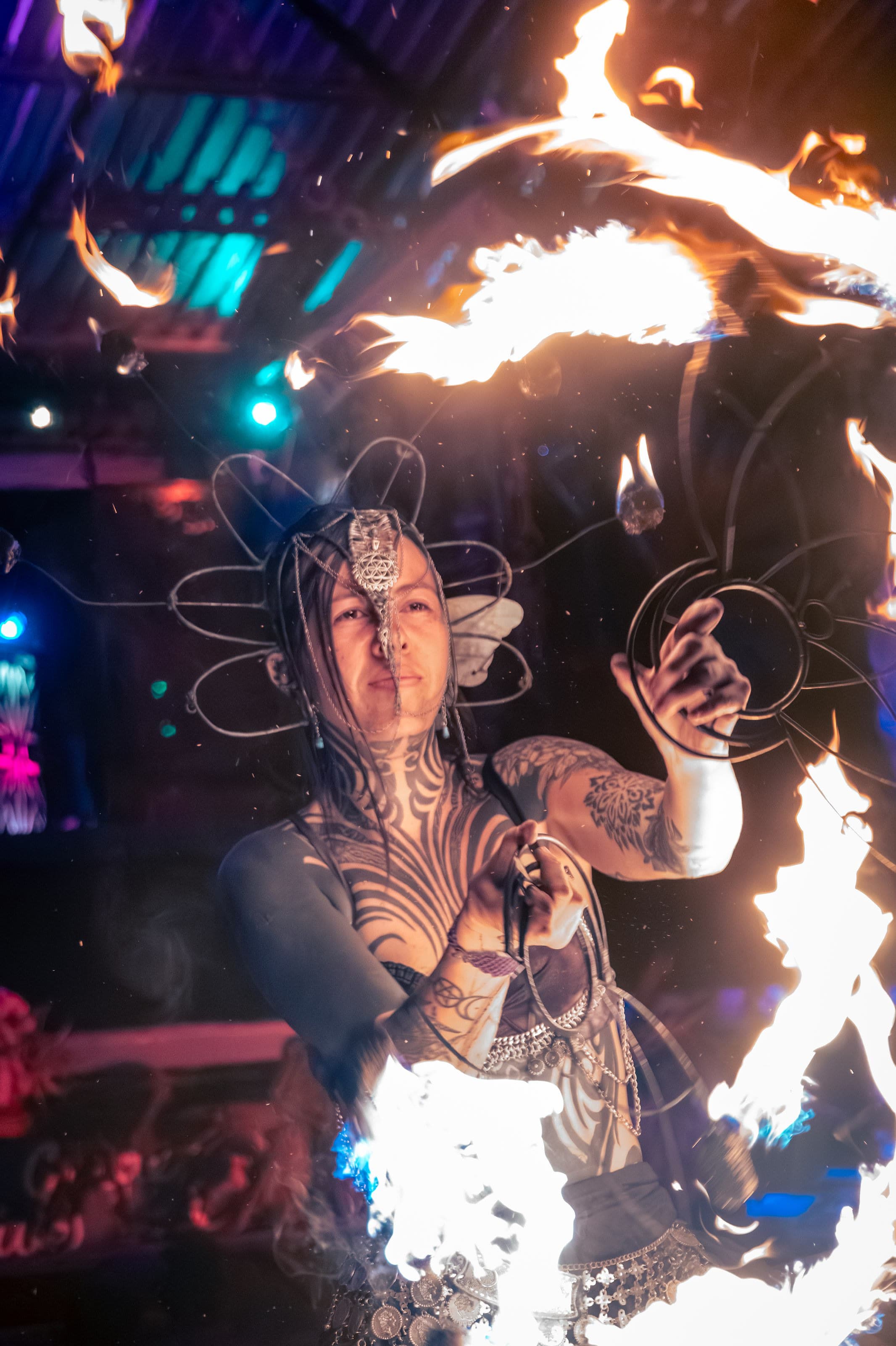 A fire performer with an ornate metal headpiece and neck tattoos, swirling orange flames around her under teal stage lights.