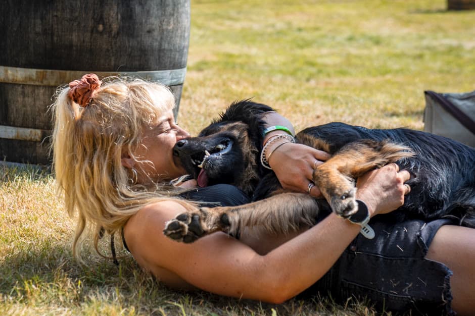 A blonde woman lies on the grass laughing as she hugs a large black-and-tan dog, a wooden barrel behind them in the sun.
