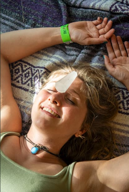A woman lies on a patterned blanket with her eyes closed and a quartz crystal on her forehead, arms open, a green wristband and a turquoise pendant.