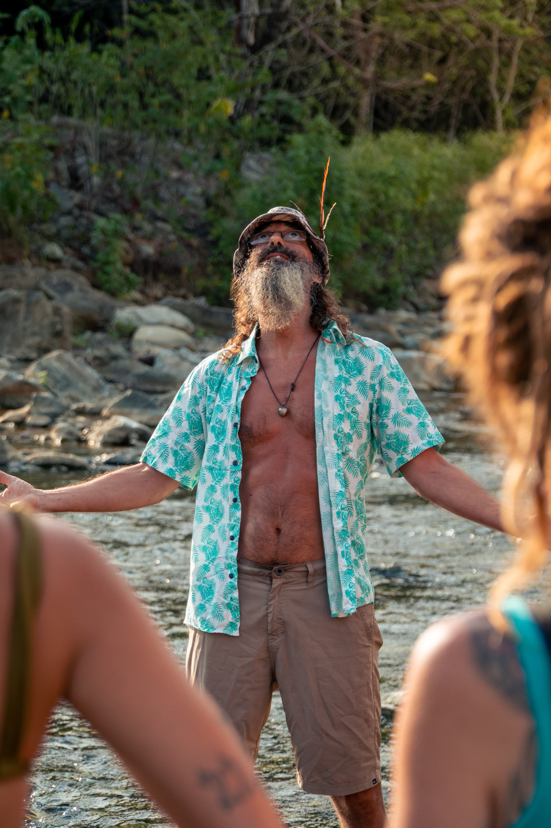 A bearded man stands in a river at golden hour with both arms outstretched, wearing a dark tank top.