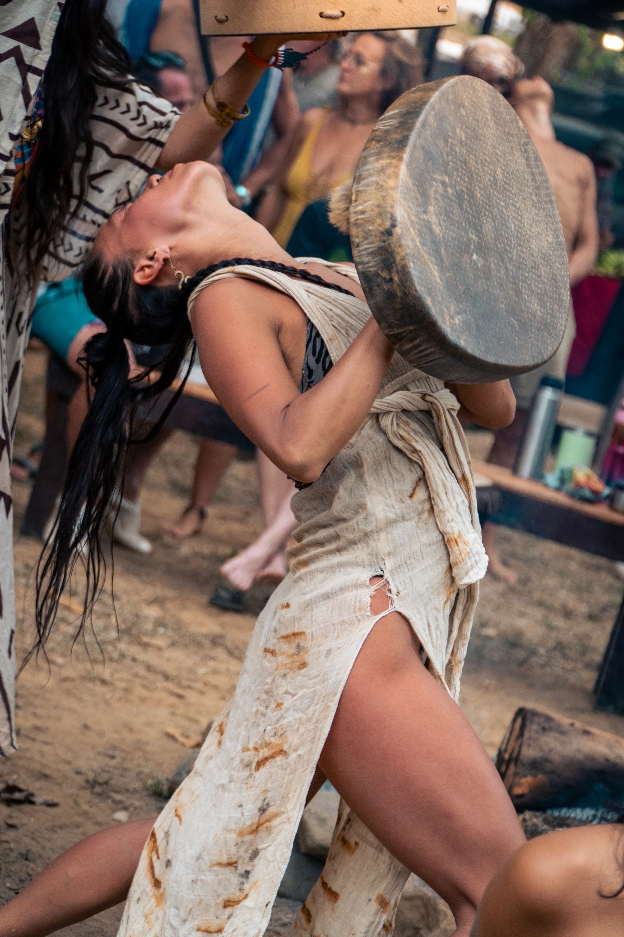 A woman leans back in an ecstatic dance, holding a large frame drum, wearing a bright patterned top and a coin belt.