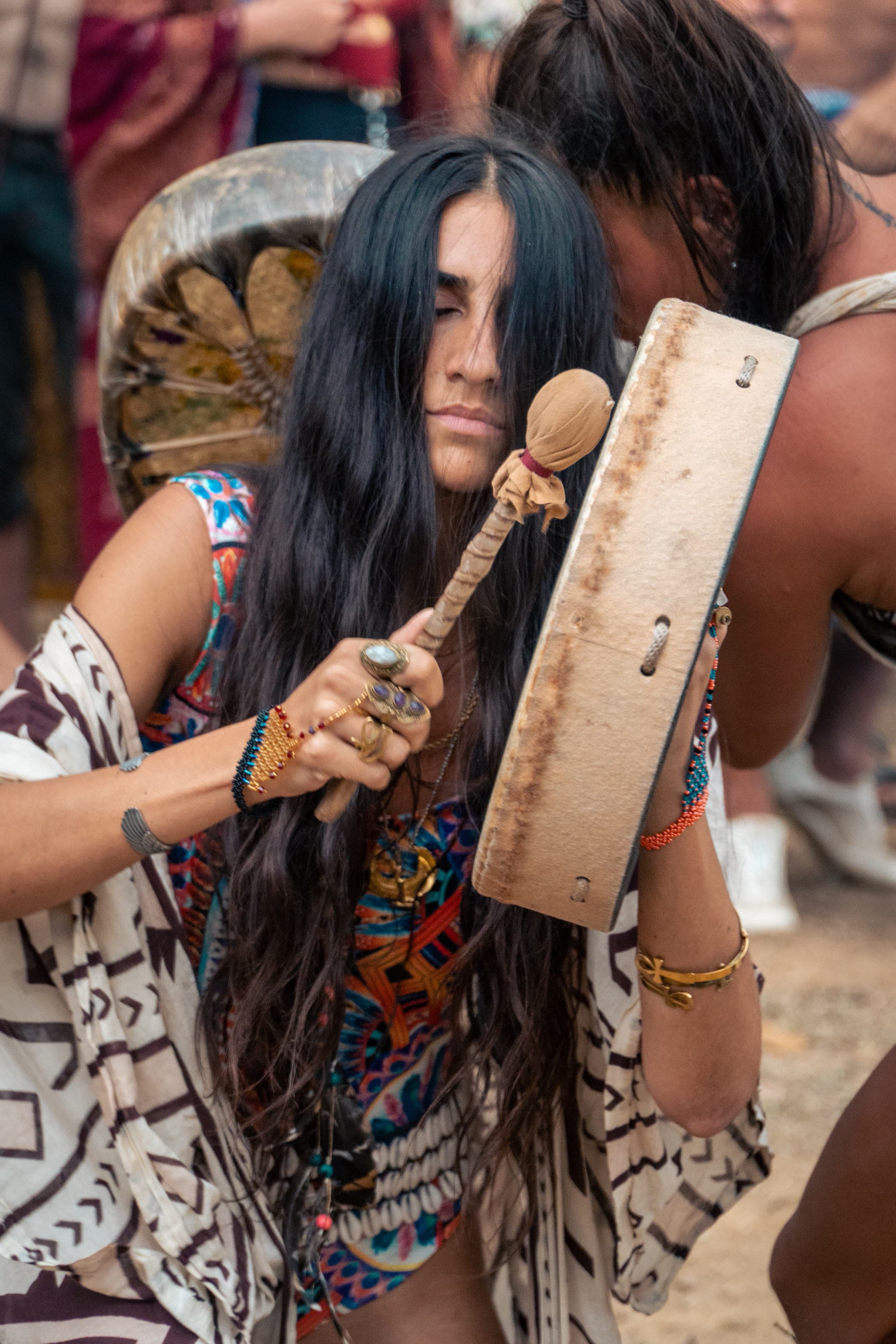 A woman plays a frame drum with her eyes closed, wearing a colorful tribal top and stacked bracelets.