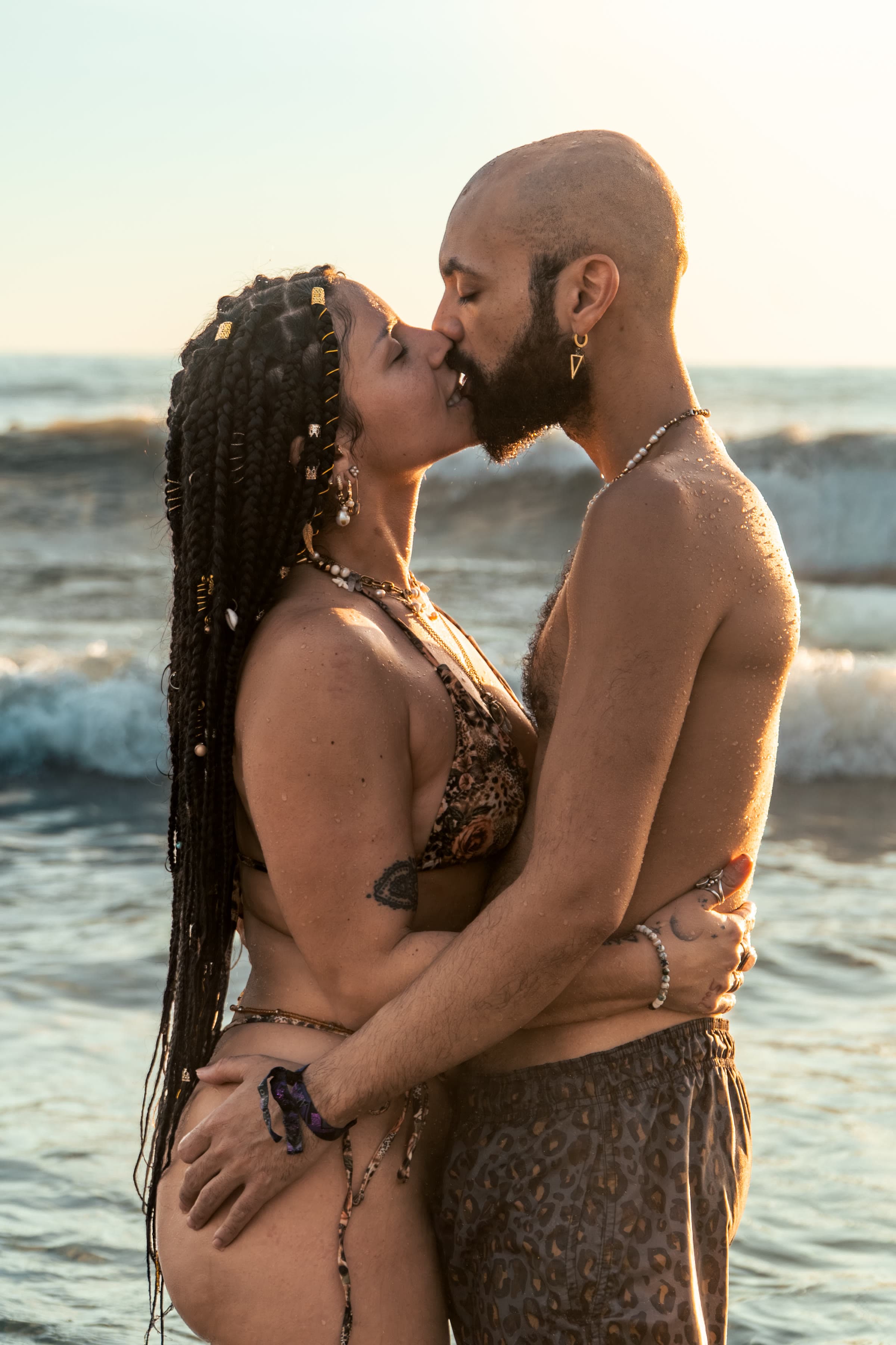 Um casal a beijar-se no oceano à hora dourada: ela com tranças escuras longas, punhos de ouro e biquíni de leopardo; ele careca e de barba, com calções de leopardo a condizer.