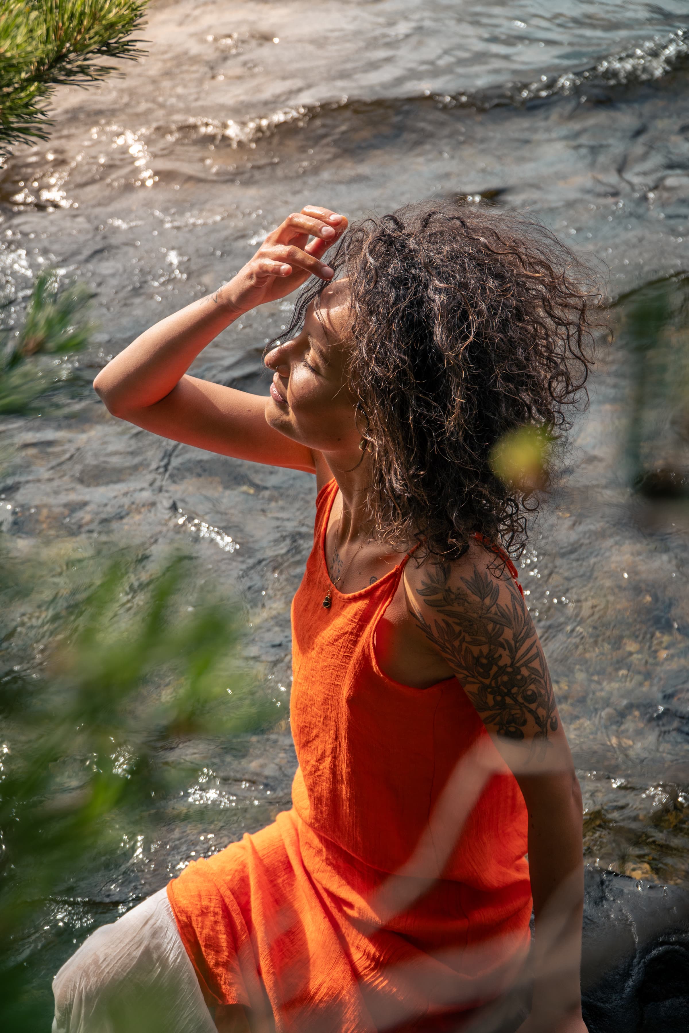 Uma mulher com o cabelo escuro molhado e os olhos fechados, de pé em água clara e pouco profunda, com um top terracota, uma tatuagem botânica no braço e agulhas de pinheiro em primeiro plano.