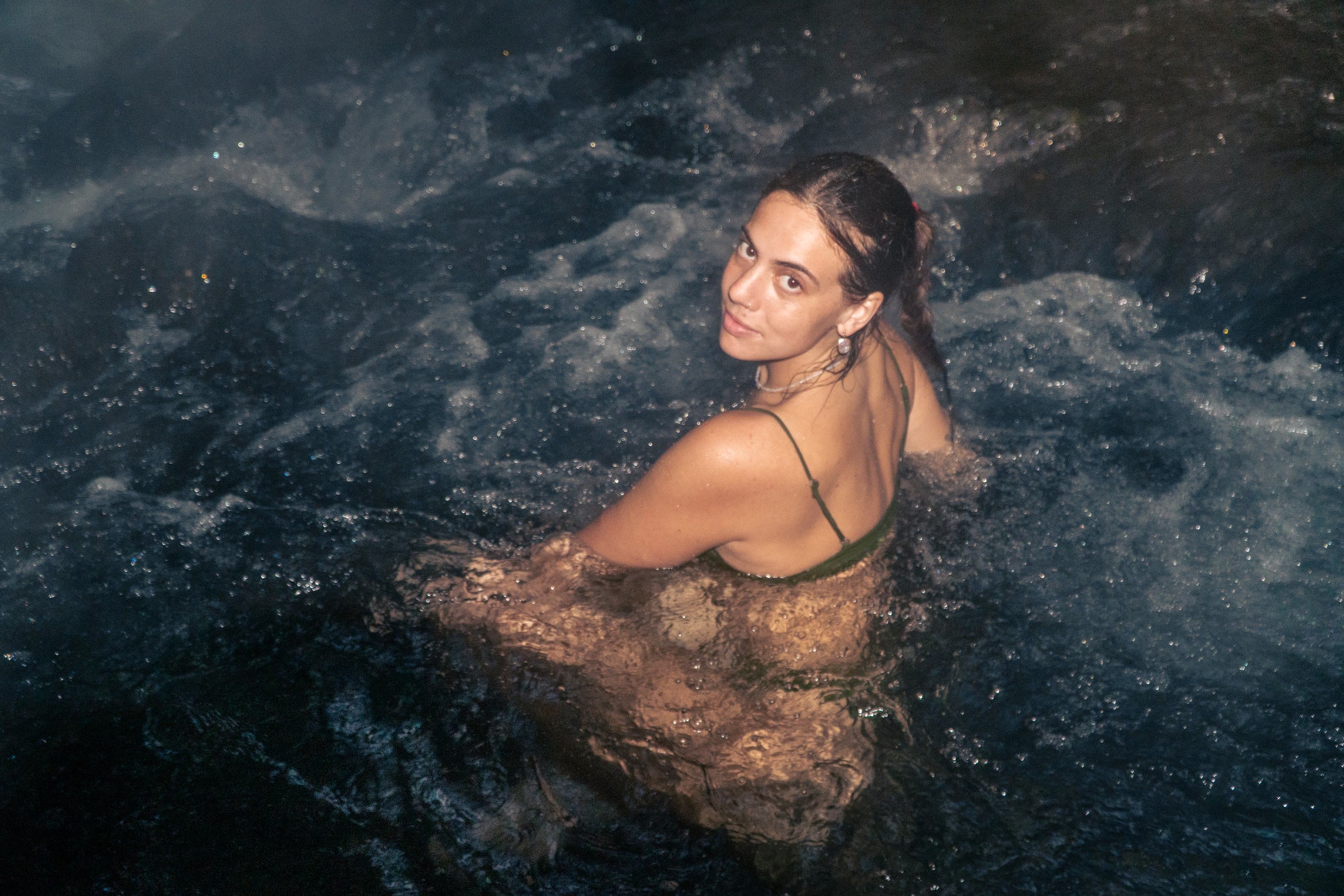 A woman with a long braid looks over her shoulder in dark bubbling water at night, wearing an olive bikini top.
