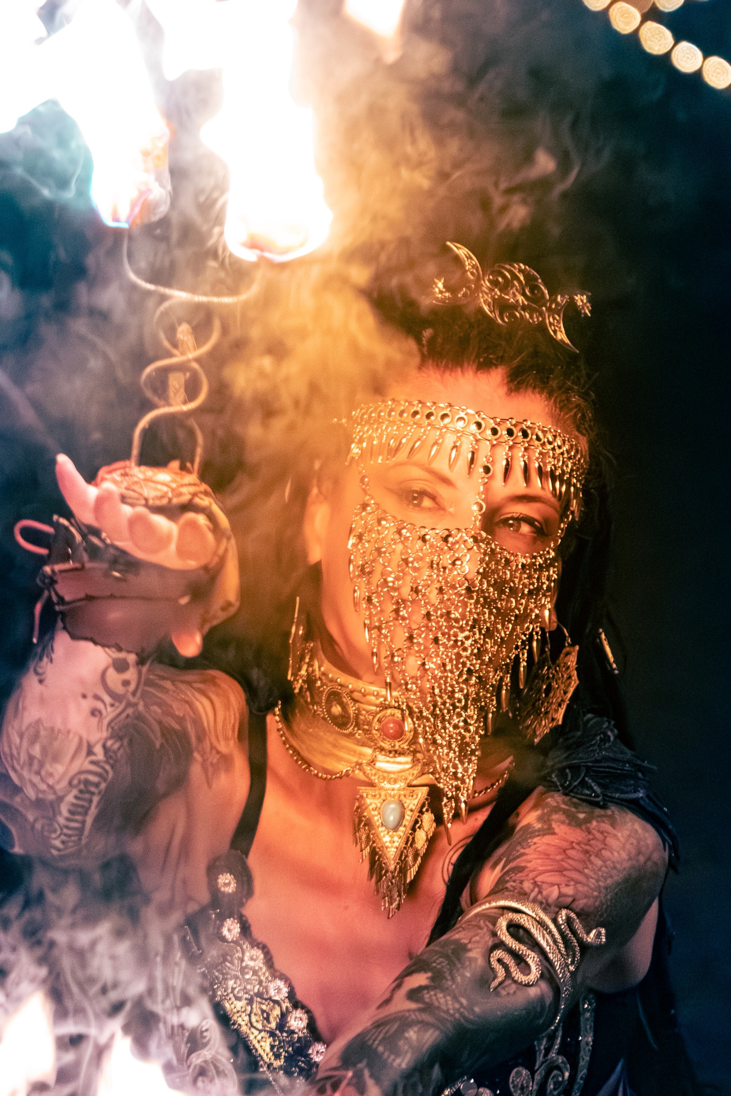 Uma artista de fogo com véu de malha dourada e adereço de lua crescente, com uma tocha em chamas entre fumo e luz quente.