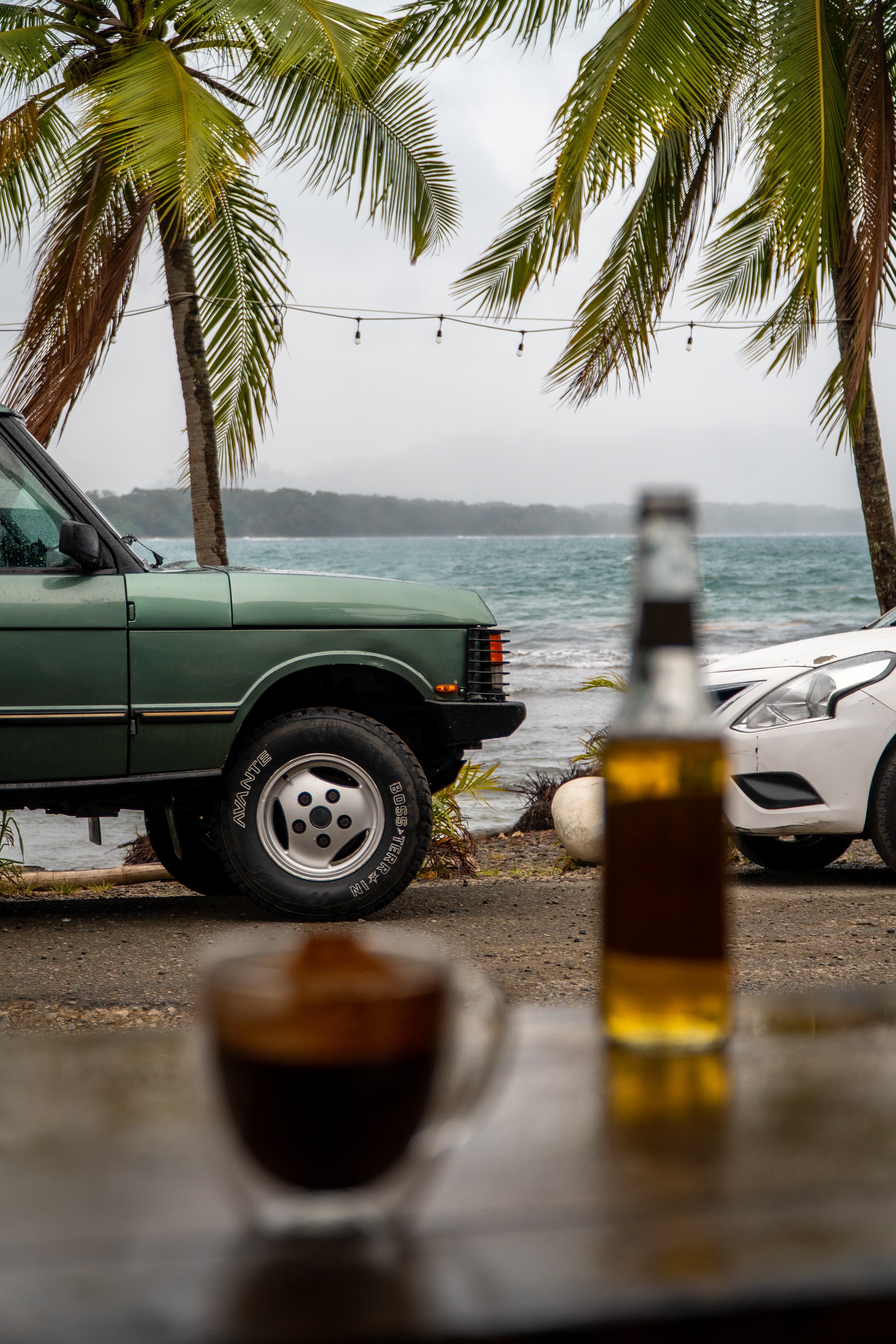 Range Rover verde-sálvia numa margem de gravilha junto a água turquesa, sob palmeiras e luzes de festão, com café e uma garrafa num parapeito de madeira em primeiro plano.
